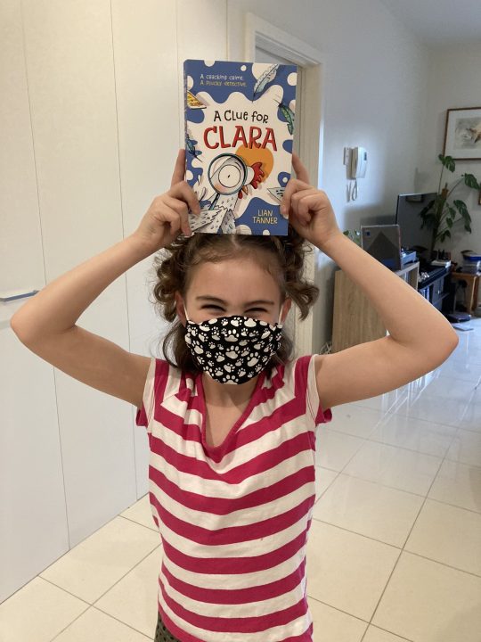 Young girl beaming above her face mask, holding copy of A Clue for Clara above her head.